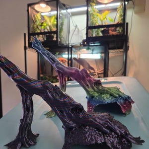 May include: Three colorful, artificial driftwood pieces, suitable for reptile habitats, are displayed on a light blue surface. The driftwood is painted in shades of purple, blue, green, and yellow. In the background, there are terrariums with plants and lighting.