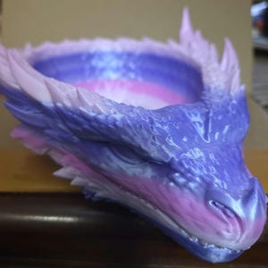May include: A 3D-printed dragon head-shaped bowl in shades of purple, pink, and white. The bowl has a detailed, textured surface with sharp, pointed features. The dragon's open mouth and detailed scales are visible.