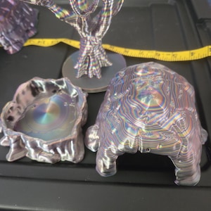 May include: A collection of iridescent, abstract sculptures. The set includes a tree-like structure, a turtle-shaped object, and a bowl-like piece. The objects have a layered, textured appearance with a metallic sheen, reflecting a spectrum of colors. The objects are on a black surface.