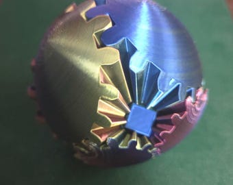 May include: A spherical object composed of interlocking gear-like segments in metallic shades of blue, gold, and pink. The structure features a central blue square component with radiating ridges that connect to the outer curved, serrated gear pieces.