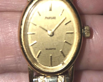 May include: A gold-tone analog wristwatch with an oval face and a textured metal link band. The watch dial features a gold background with thin gold hour markers, gold hands, and the word Pulsar printed near the top with Quartz at the bottom.
