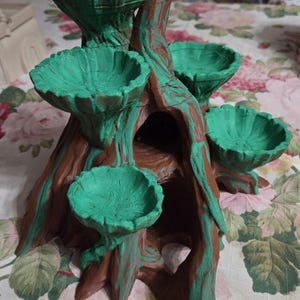 May include: A brown and green tree-shaped display stand featuring four bowl-like platforms extending from a central trunk. The structure has a textured bark-like finish with green accents on the upper surfaces of the bowls and along the trunk.