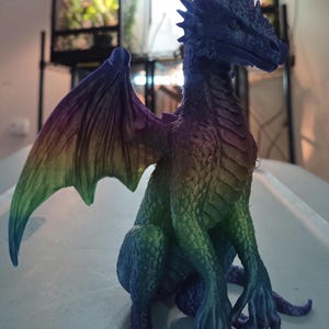 May include: A colorful dragon figurine with a rainbow gradient on its wings and body. The dragon is in a seated position, with its wings spread and its head held high. The figurine is made of a textured material and has a detailed design.