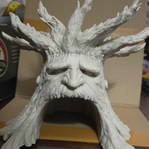 May include: A light gray, detailed sculpture of a tree with a face. The tree has a large, open mouth and a long, flowing beard. The sculpture's face has a sad expression, with furrowed brows and downturned lips. The tree's branches extend upwards.