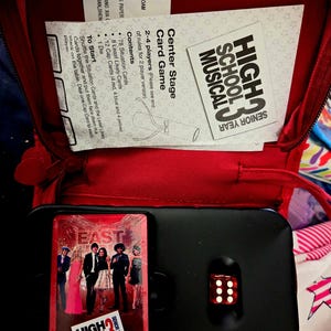 May include: A red zippered case containing a High School Musical 3 card game. The game includes cards, a die, and instructions. The card game features images of the cast and the text "High School Musical 3 Senior Year".