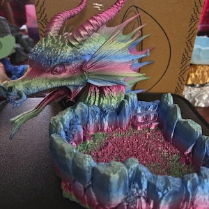 May include: A colorful dragon sculpture with a detailed head, horns, and wings. The dragon's head is a mix of pink, blue, and green hues. It sits above a rocky, multi-colored base, creating a fantasy-themed decorative piece.