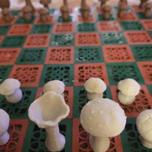 May include: A unique chess set featuring mushroom-shaped pieces in shades of brown and white. The board has a red and green checkered pattern. The pieces are arranged on the board, ready for play.