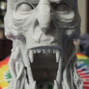 May include: A detailed, light gray 3D-printed sculpture of a monster's head with an open mouth, sharp teeth, and intricate facial features. The sculpture has a textured surface and is set against a colorful background.