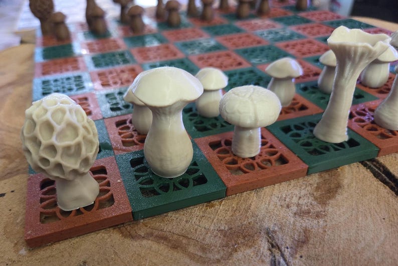 May include: A unique mushroom-themed chess set. The chess pieces are white mushroom figures, set on a board with red and green squares. The board has a decorative, openwork design. The pieces include morels and other mushroom shapes.