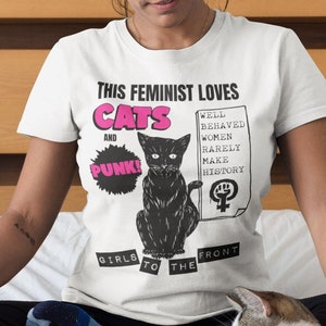 May include: White t-shirt with a black and white illustration of a cat and the text "This Feminist Loves Cats and Punk!"  The text "Well Behaved Women Rarely Make History" is written on a white rectangle with a pink feminist symbol. The text "Girls to the Front" is written on a black banner.
