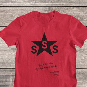 May include: Red t-shirt with a black star featuring the letter "S" three times. The text "Lips Lips Guns Guitar Hey! Jayne Mansfield Superstar" is printed below the star.
