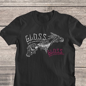 May include: Black t-shirt featuring the text "G.L.O.S.S." in white and pink, with the tagline "(Girls Living Outside Society's Shit)". The graphic includes a stylized, skeletal dragon design. The shirt is made of a soft material.