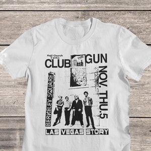 Può includere: Una maglietta bianca con una stampa grafica in bianco e nero di una band chiamata "The Gun Club". La grafica include il nome della band, la data "Nov. Thu. 5", e il testo "Las Vegas Story".