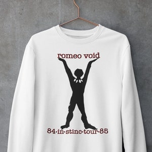 May include: White sweatshirt featuring a black silhouette figure with arms raised, holding the words "romeo void" above its head. Below the figure is the text "84-in-stinc-tour-85" in a maroon color. The sweatshirt is hanging on a copper-colored hanger.