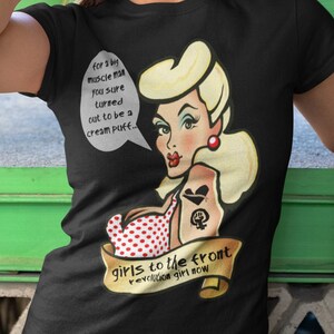 May include: Black t-shirt with a cartoon illustration of a woman with blonde hair and red polka dot swimsuit. The text on the shirt reads "for a big muscle man you sure turned out to be a cream puff... girls to the front revolution girl now"