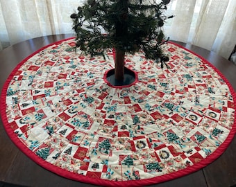 May include: A round Christmas tree skirt with a red border and a patchwork design. The patchwork features squares with Christmas trees, wreaths, and other holiday motifs in red, green, and cream colors. The skirt is placed around a small Christmas tree.
