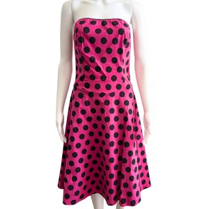 Puede incluir: Vestido sin tirantes, hasta la rodilla, en un vibrante color rosa, adornado con grandes lunares negros espaciados uniformemente. El vestido presenta un corpiño ajustado y una falda acampanada, creando una silueta clásica. El borde superior está adornado con encaje negro.