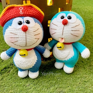 May include: Two crocheted stuffed toys of a popular Japanese cartoon character, Doraemon. One is wearing a red and yellow hat, the other is blue. Both have white faces with black eyes and a yellow bell around their necks.