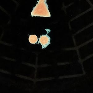 May include: A black surface features a glowing light blue spider web pattern. Above the web, there is a peach-coloured hourglass shape and two small peach-coloured circles, all outlined with a glowing light blue border against the dark background.