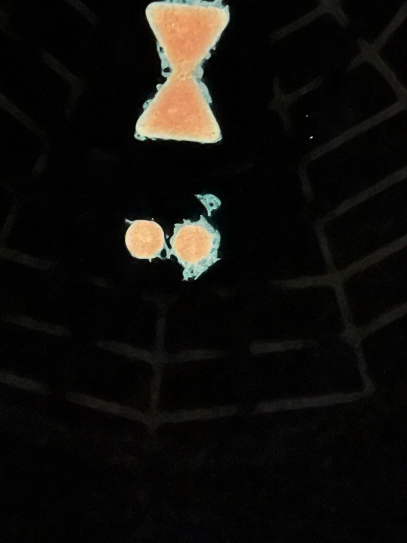 May include: A black surface features a glowing light blue spider web pattern. Above the web, there is a peach-coloured hourglass shape and two small peach-coloured circles, all outlined with a glowing light blue border against the dark background.
