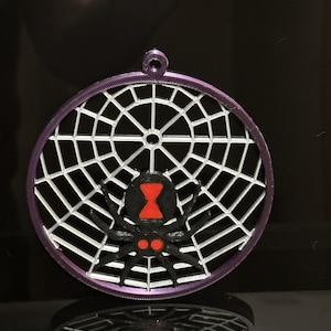 May include: A circular purple ornament featuring a white spider web pattern and a black spider with a red hourglass marking on its abdomen and two red spots near its head. The ornament has a small loop at the top for hanging purposes.