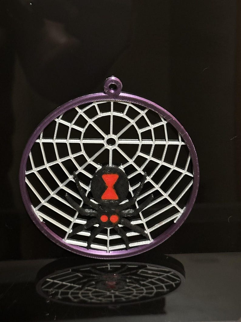 May include: A circular purple ornament featuring a white spider web pattern and a black spider with a red hourglass marking on its abdomen and two red spots near its head. The ornament has a small loop at the top for hanging purposes.