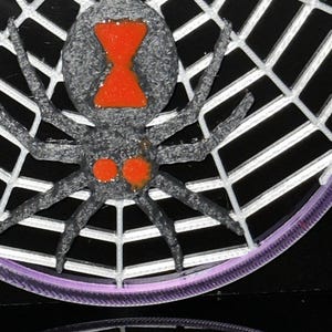 May include: A three-dimensional black spider with a textured surface and a bright orange hourglass marking on its abdomen, plus two orange spots near the head, positioned on a white web structure with a purple circular border at the base.