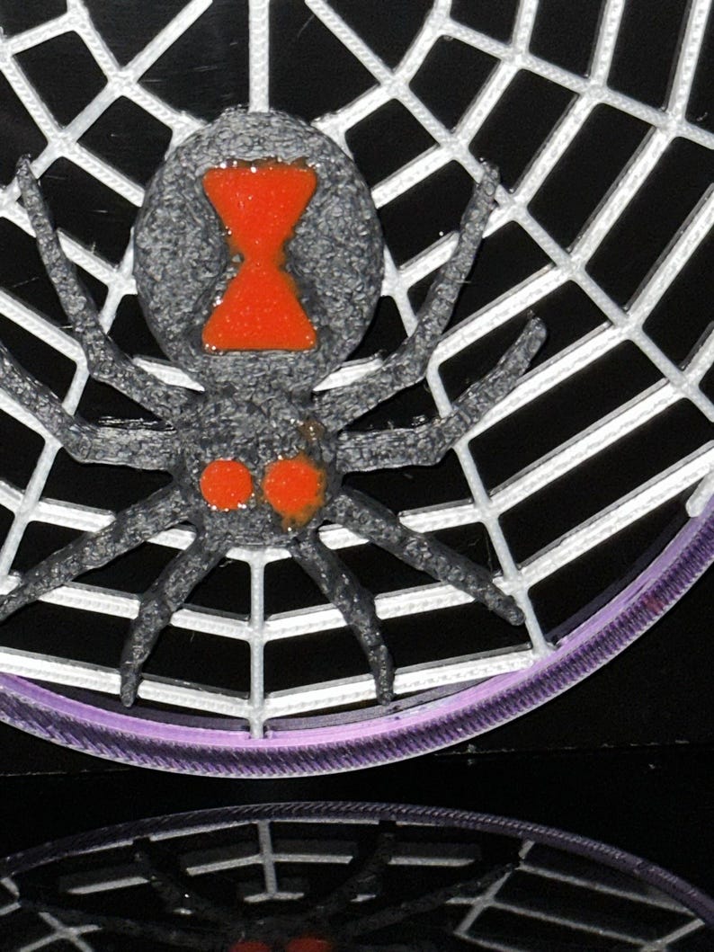 May include: A three-dimensional black spider with a textured surface and a bright orange hourglass marking on its abdomen, plus two orange spots near the head, positioned on a white web structure with a purple circular border at the base.