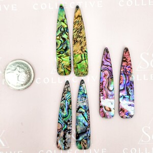 May include: Six elongated, iridescent abalone shell pendants in shades of green, blue, purple, and gold. A silver coin is next to the pendants for size comparison. The pendants have a pointed top and a flat bottom.