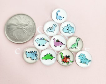 Puede incluir: Diez cabujones redondos de vidrio blanco, cada uno con una ilustración diferente de dinosaurio de dibujos animados. Los dinosaurios son azules, verdes y rosas. Una moneda canadiense está al lado de los cabujones para comparar el tamaño. Los cabujones miden unos 2,5 cm de diámetro.