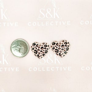 May include: Two heart-shaped earrings with a leopard print pattern, next to a coin. The earrings have a light pink background with black and brown spots. The earrings are likely for pierced ears.