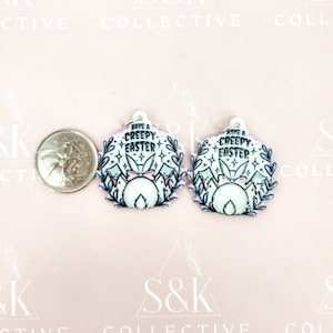 May include: Two Easter-themed earrings in white and pastel colours, each with the text "Have a Creepy Easter". The earrings feature a bunny tail design with a pink bow, surrounded by a floral wreath. A coin is next to the earrings.