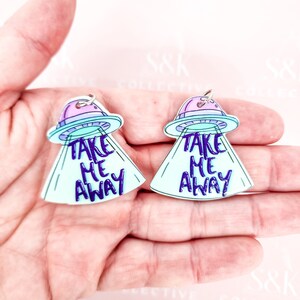May include: Two alien spaceship charms with the text "TAKE ME AWAY" in purple. The spaceships have a light blue beam and a purple and light blue top. The charms are held in a hand.