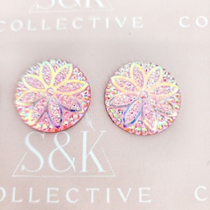 May include: Two iridescent pink flower-shaped stud earrings. Each earring features a floral design with a central flower and surrounding petals, encrusted with sparkling crystals. The earrings are round and have a light pink background.