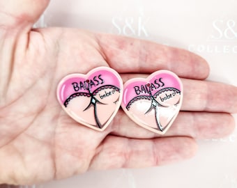 Puede incluir: Dos broches en forma de corazón con un diseño rosa y color carne. Cada broche presenta la palabra "BADASS" en negro, con un dibujo de un trasero y la palabra "babe" debajo. Los broches se sostienen en una mano.