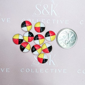May include: A group of round cabochons with a four-part design in red, black, yellow, and white. A coin is next to the cabochons for size comparison. The background is a light pink surface with the text "S&K COLLECTIVE".