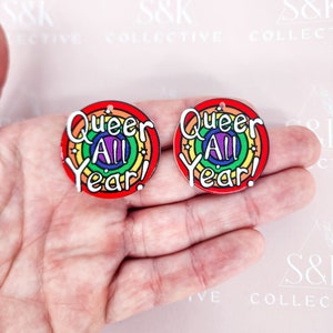 Puede incluir: Dos pendientes redondos y coloridos con un diseño de arcoíris y las palabras "Queer All Year!" en blanco. Los pendientes tienen un borde rojo y un pequeño agujero en la parte superior para colgar. El fondo es rosa claro.