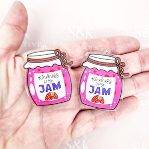 May include: Two clear acrylic charms shaped like jam jars. The jars are pink with red polka dots and a white label that reads "Kindness is my JAM." Each jar has a red bow and a strawberry illustration.