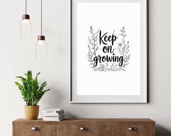 Puede incluir: Un cuadro enmarcado con la frase "Keep on growing" en escritura negra, rodeada de ilustraciones botánicas. La impresión se muestra sobre un mueble de madera con una planta en maceta e iluminación decorativa.