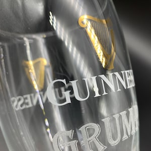 Puede incluir: Un vaso de pinta de vidrio transparente con un logotipo de arpa dorado y la palabra "GUINNESS" grabada en blanco. La parte inferior del vaso tiene la palabra "GRUM" grabada en blanco.