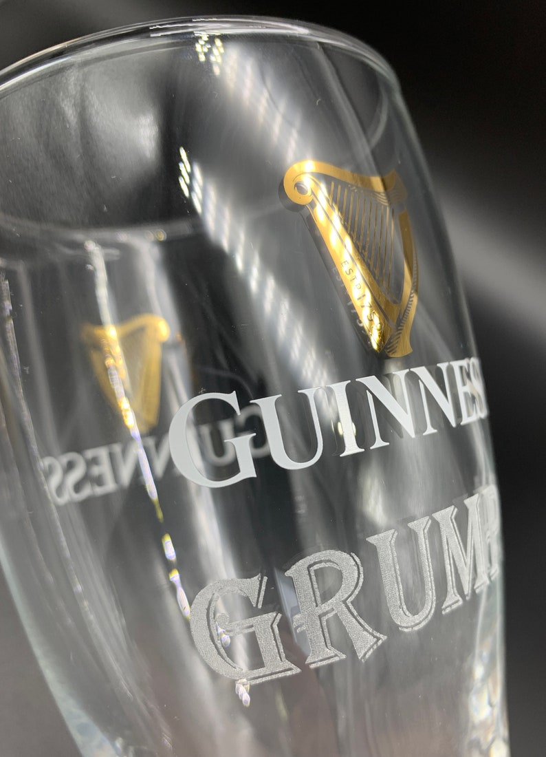 Puede incluir: Un vaso de pinta de vidrio transparente con un logotipo de arpa dorado y la palabra "GUINNESS" grabada en blanco. La parte inferior del vaso tiene la palabra "GRUM" grabada en blanco.