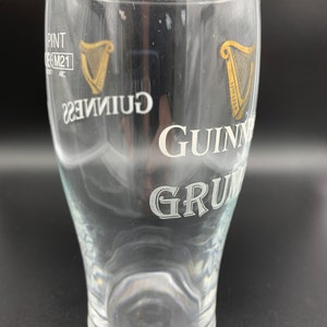 Puede incluir: Un vaso de pinta de vidrio transparente con un logotipo de Guinness blanco y un logotipo de arpa dorado. El vaso est&aacute; vac&iacute;o.
