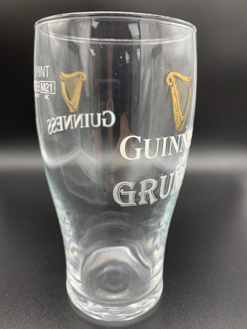 Puede incluir: Un vaso de pinta de vidrio transparente con un logotipo de Guinness blanco y un logotipo de arpa dorado. El vaso est&aacute; vac&iacute;o.