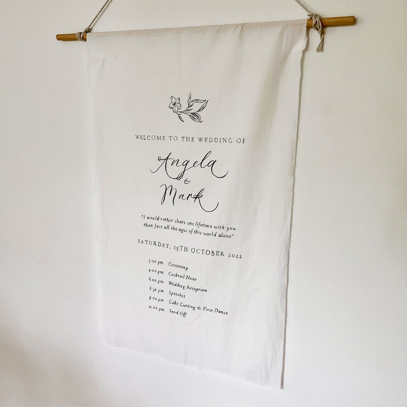 May include: A white canvas wedding banner with black text announcing the wedding of Angela and Mark. The banner includes the date, time, and a list of events for the wedding day. The text reads: "Welcome to the wedding of Angela & Mark. 'I would rather share one lifetime with you than face all the ages of this world alone.' Saturday, 15th October 2022. 3:00 pm Ceremony. 4:00 pm Cocktail Hour. 6:00 pm Wedding Reception. 8:30 pm Speeches. 9:00 pm Cake Cutting & First Dance. 11:00 pm Send Off."