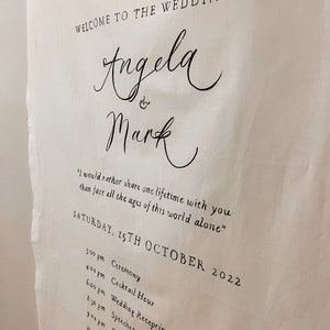 May include: A white wedding sign with black text announcing the wedding of Angela and Mark. The sign includes the date, Saturday, 15th October 2022, and a list of events with times.