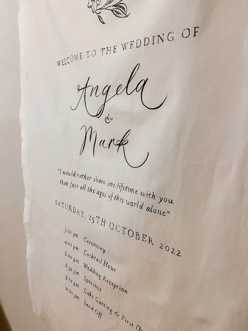 May include: A white wedding sign with black text announcing the wedding of Angela and Mark. The sign includes the date, Saturday, 15th October 2022, and a list of events with times.