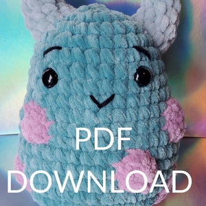 May include: A crocheted plush toy in the shape of a blue monster with pink cheeks. The toy has black eyes and a stitched-on smile. The text "PDF DOWNLOAD" is printed below the toy. The text "*** NOT A PHYSICAL ITEM ***" is printed below the text.