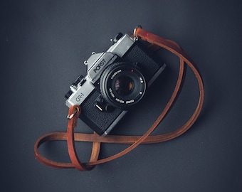 May include: A silver and black 35mm film camera with a brown leather strap. The camera has a lens with the text "PORST COLOR" and "1:1.9/50 mm" printed on it. The camera is on a dark grey background.