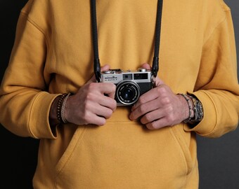 May include: A person wearing a yellow hoodie with a black camera strap and a silver camera with the text "minolta hi-matic 7" hanging around their neck. The person is wearing a brown bracelet on their left wrist and a silver watch on their right wrist.