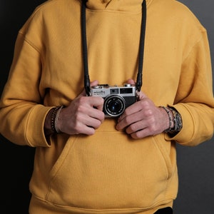 Pode incluir: Uma pessoa vestindo um moletom amarelo com uma alça de câmera preta e uma câmera prateada com o texto "minolta hi-matic 7" pendurada no pescoço. A pessoa está usando uma pulseira marrom no pulso esquerdo e um relógio prateado no pulso direito.